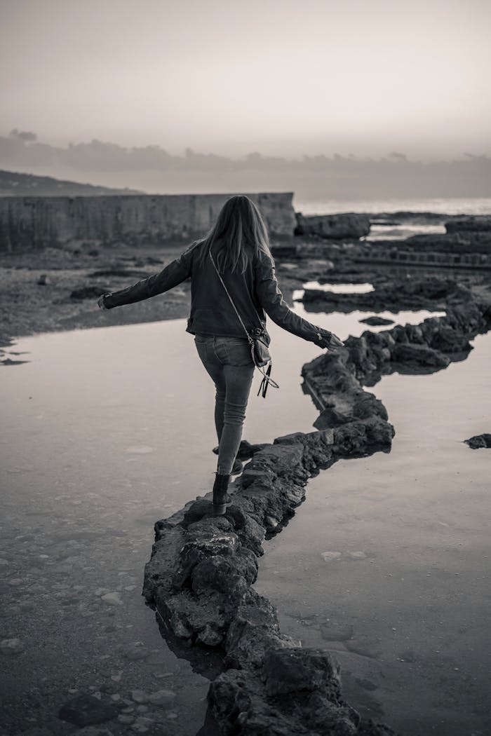 gallery-4 A woman walks along a rocky path by the water at twilight, exuding a sense of adventure and solitude.